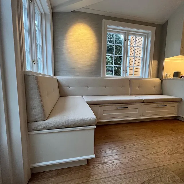 Innebygd vindusbenk med skuffer under, lyst trekk og puter, plassert i en lun krok ved et vindu med naturlig lys.