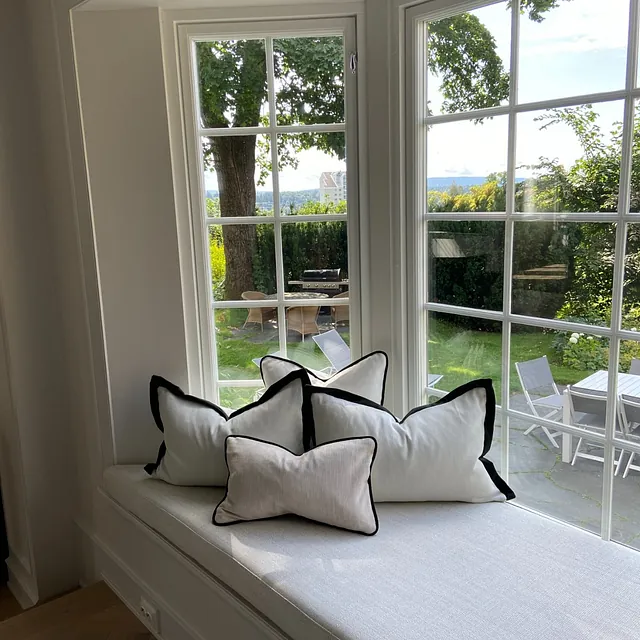 Daybed med lyse puter i moderne bolig, plassert foran stort vindu med utsikt mot hage.