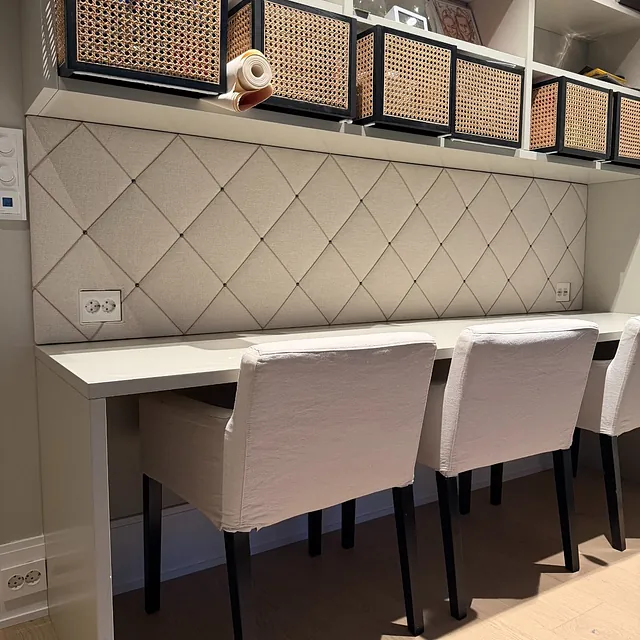 Arbeidsplass med polstret veggpanel i rutemønster, hyller over og stoler ved skrivebord.