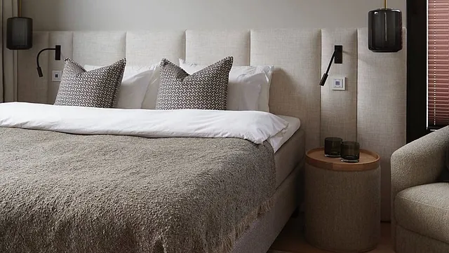 Hotellseng med polstret sengegavl, puter og nattbord i elegant og dempet soverom.