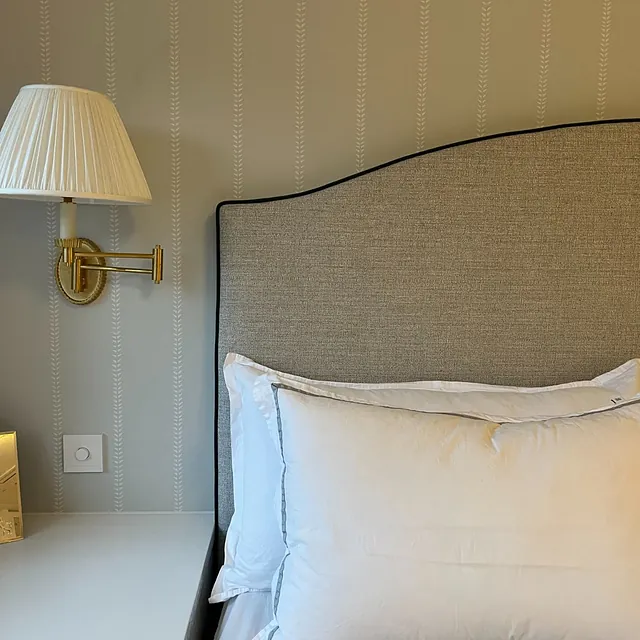 Seng med polstret sengegavl i lyst stoff, hvitt sengetøy og vegglampe montert over nattbord.