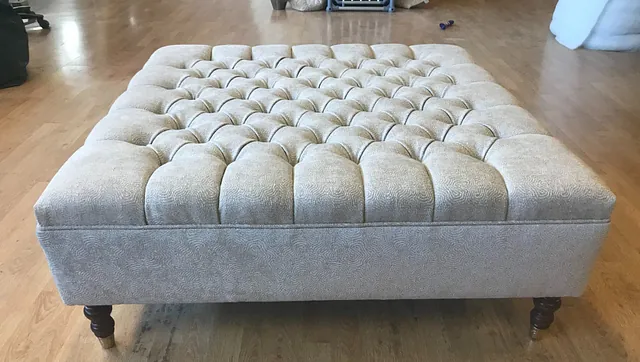 Stor kvadratisk puff med knappet polstring i lyst stoff, stående på lave ben på tregulv.